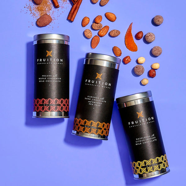 Three metal tins with black labels on a blue background, with a variety of chocolate nuts and spices spilling out. The labels on the tins read "fruition chocolate works: pecans with maple cinnamon milk chocolate, almonds with dark chocolate and smoked sea salt, and hazelnuts with citrus infused milk chocolate"