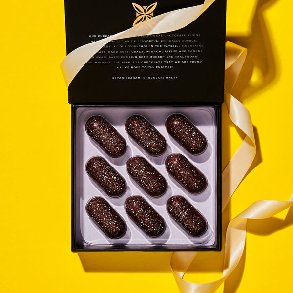 black and white confection box, open and wrapped with a cream color ribbon on a yellow background. visible inside the box are 9 oval shaped chocolate confections with copper and white color sprinkled over it