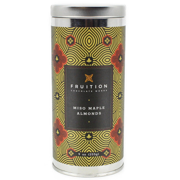 A silver tin with a black, gold, and red ornate label that reads "fruition chocolate works, miso maple almonds"