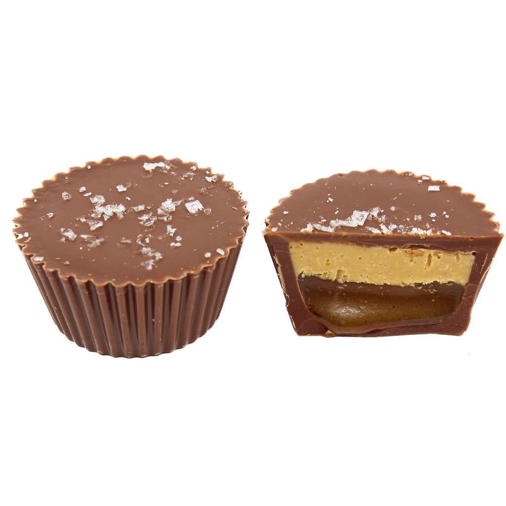 one whole peanut butter cup sitting next to one cut in half, displaying a layer of oozing caramel and a layer of peanut butter. The cups are topped with salt