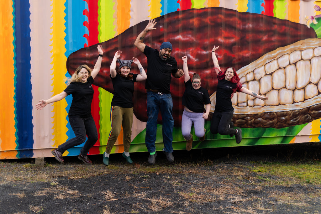 five people jumping in the air facing the camera, all wearing black shirts that say fruition chocolate works on the front