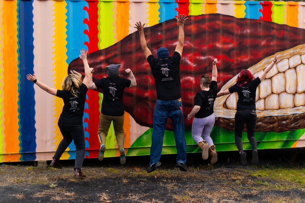 five people jumping in the air with their backs to the camera, all wearing black shirts that say Fruition Chocolate Works with a cocoa pod logo