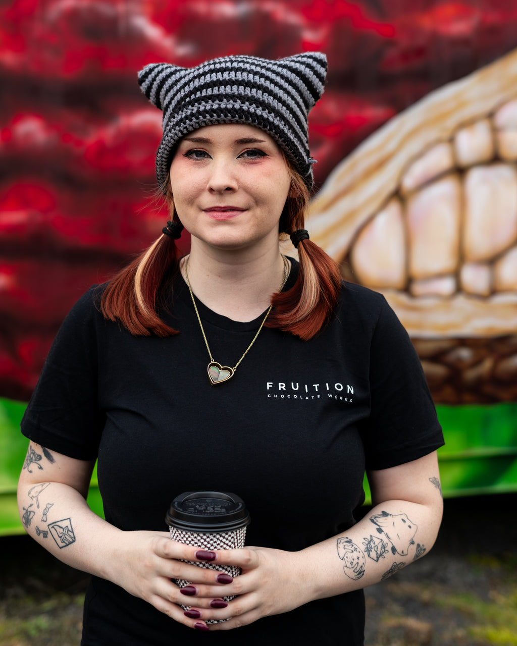girl wearing a hat and holding a coffee cup, wearing a black t-shirt that says "fruition chocolate works" on the front