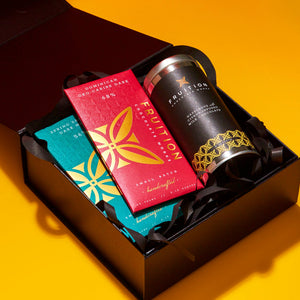 Black gift box on a yellow background containing three products: a red chocolate bar called dominican oko caribe dark 68%, a green chocolate bar called spring salted dark milk 56%, and a tin of hazelnuts with citrus infused milk chocolate