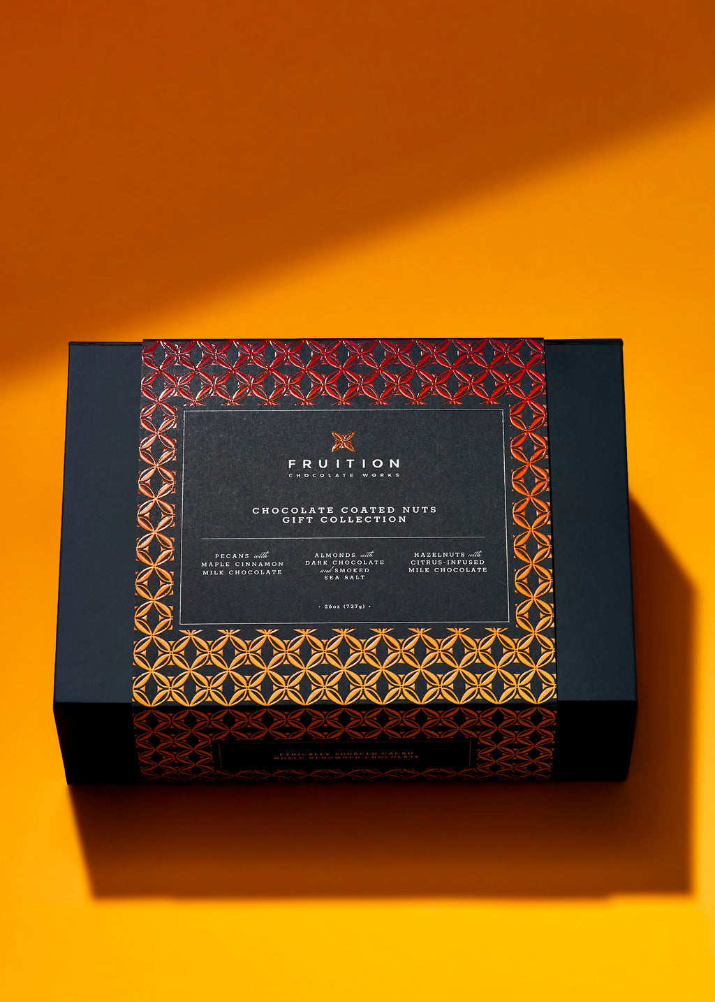 Black matte gift box with a black, yellow,  and red gradient belly band that reads "fruition chocolate: chocolate coated nuts gift collection". The box sits on an orange background