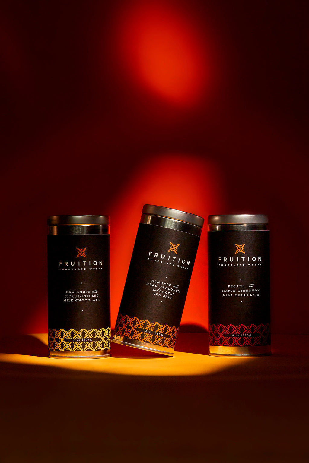 Three metal tins with black labels on a red and orange background, with dramatic lighting on them. the tins read "fruition chocolate works: hazelnuts with citrus infused milk chocolate, almonds with dark chocolate and smoked sea salt, and pecans with maple cinnamon milk chocolate"