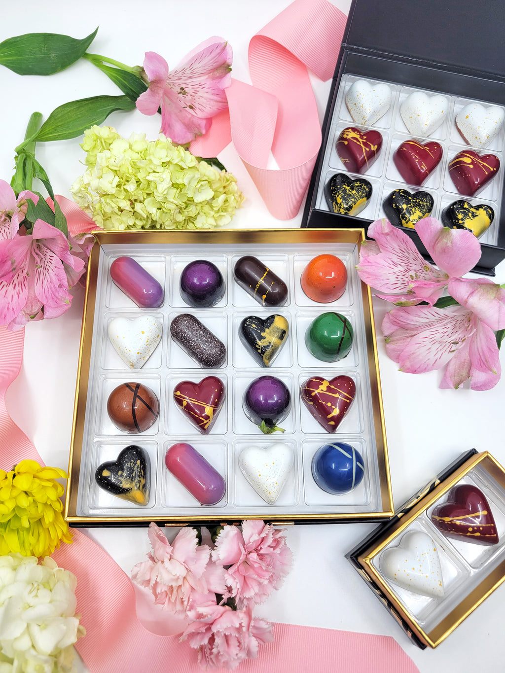 Black confection box containing 16 chocolate confections of various shapes and colors, surrounded by pink and yellow flowers- Fruition Chocolate