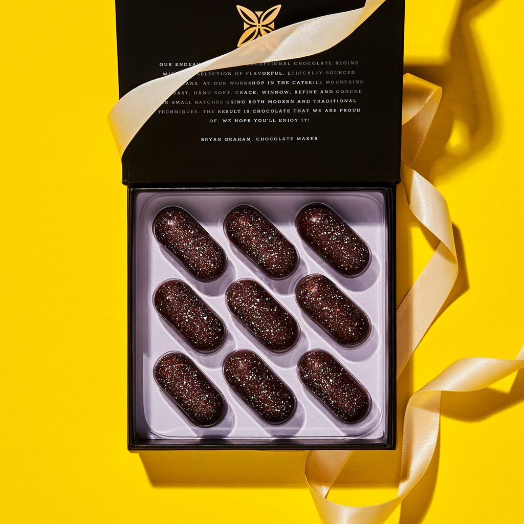 black and white confection box, open and wrapped with a cream color ribbon on a yellow background. visible inside the box are 9 oval shaped chocolate confections with copper and white color sprinkled over it