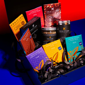 Black vinyl gift box containing 7 various chocolate bars, a confection box, a jar of marcona almonds, and a tin of pecans. The box is stuffed with black paper spiral fill, and placed on a dark blue and red background