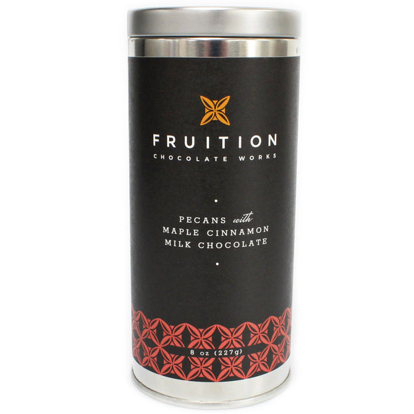 tall narrow metal tin with a black label with reddish pattern along the bottom. The label reads "fruition chocolate works: pecans with maple cinnamon milk chocolate"