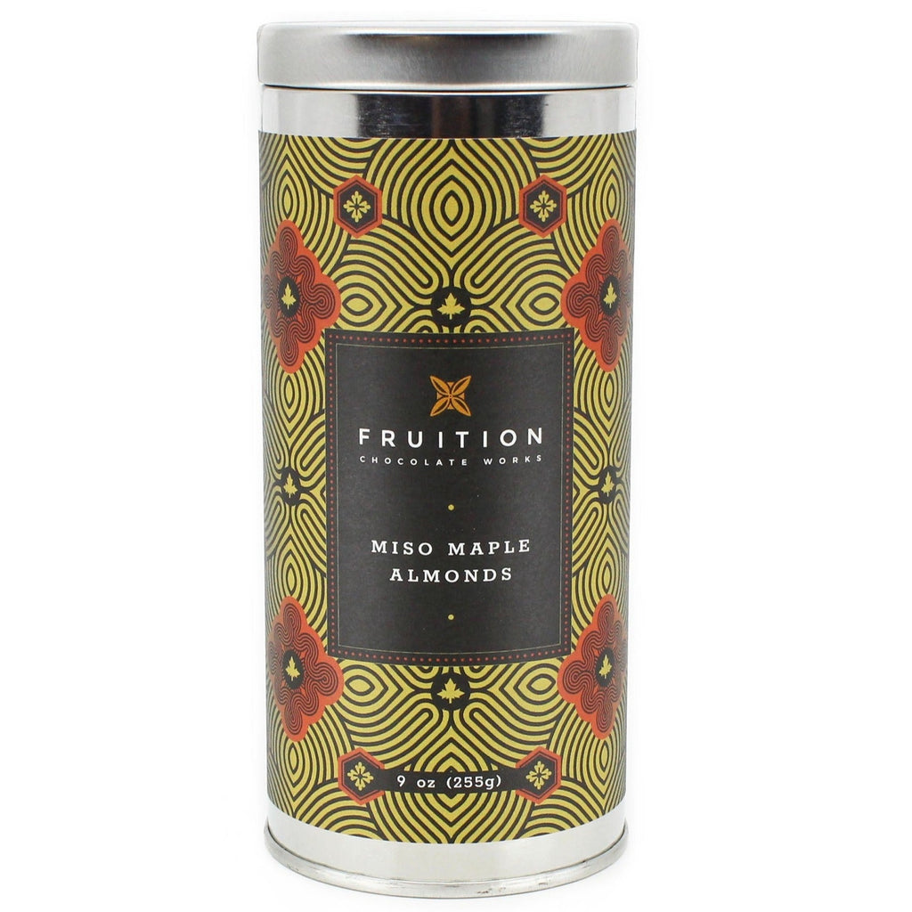 A silver tin with a black, gold, and red ornate label that reads "fruition chocolate works, miso maple almonds"